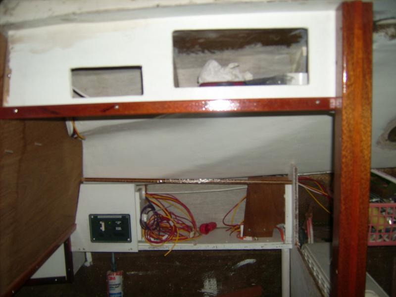 2 March 2012.  Overhead panel, above the quarter berth.  In the after (smaller) hole goes the VHF; above it go the horn switch, deck-outlet switch and cabin footlights switch.  The bigger hole is the all-too-necessary wallets-and-sunglasses locker.  In the opening outboard is the electrical panel; the drop-down door is removed for painting but the 115vac panel is there.  I installed this bulkhead to divide the quarter berth from the settee; the vertical post is both to grab (thus precluding the need for overhead handrails) and to stiffen the end of the bulkhead.