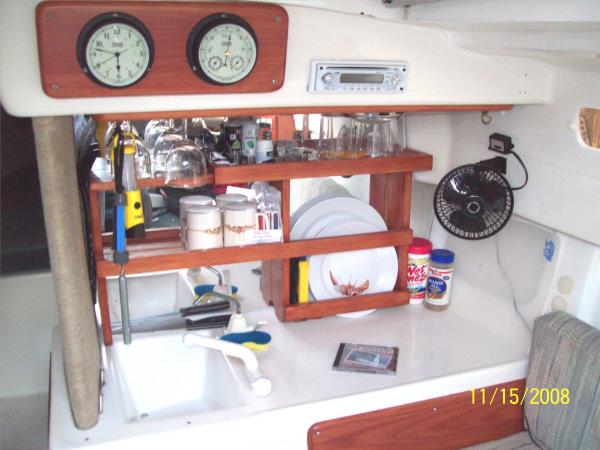 Galley dish/glass rack. Clock and barometer.  AM/FM/CD. Fan.  Front faucet is pressurized from 13-gal. tank.  Original pump tap (rear) is used as a wine dispenser.  Cheap Port of course.  After all, any port in a storm. There is a 12V outlet under the fan.  The grey wire is another fan plugged in.