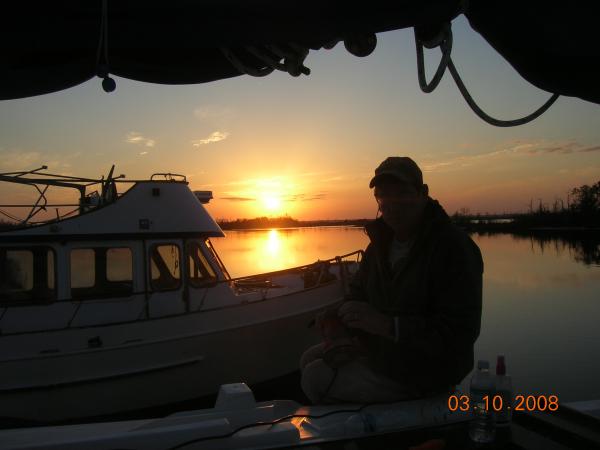 Mike R. sitting in the cockpit at sundown while the boat is on the hard during our exterior overhaul (summer 2008). The background is the mouth of the Escatawpa River at it's confluence with the Pascagoula River.